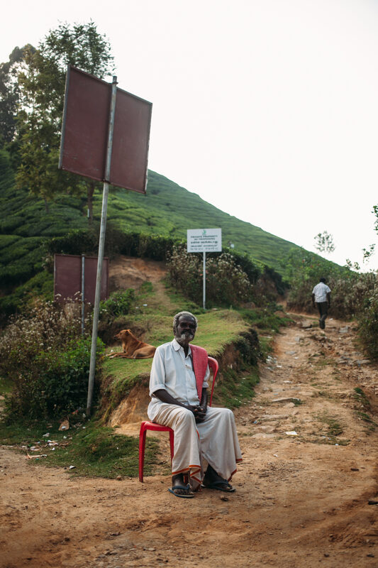 A blind man sits quietly on a red chair beside a Munnar hill road, listening to the world around him. print option
