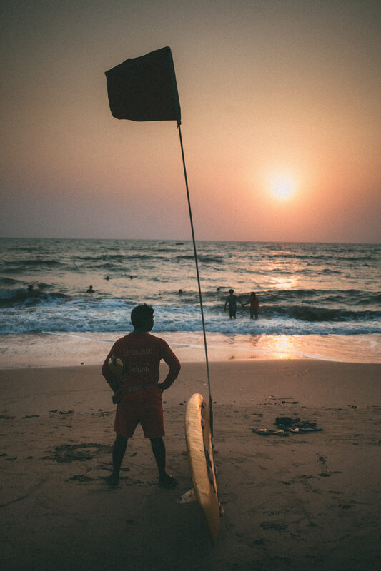 A Goa beach lifeguard board and safety flag turn into a sunset silhouette above the surf. print option