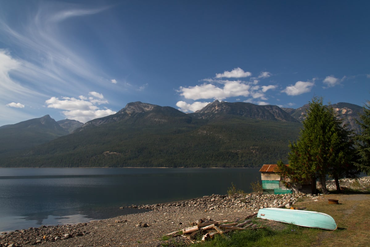 A green canoe rests on a rocky shore below mountain water and sky in the Kootenays near Kaslo, BC. print option