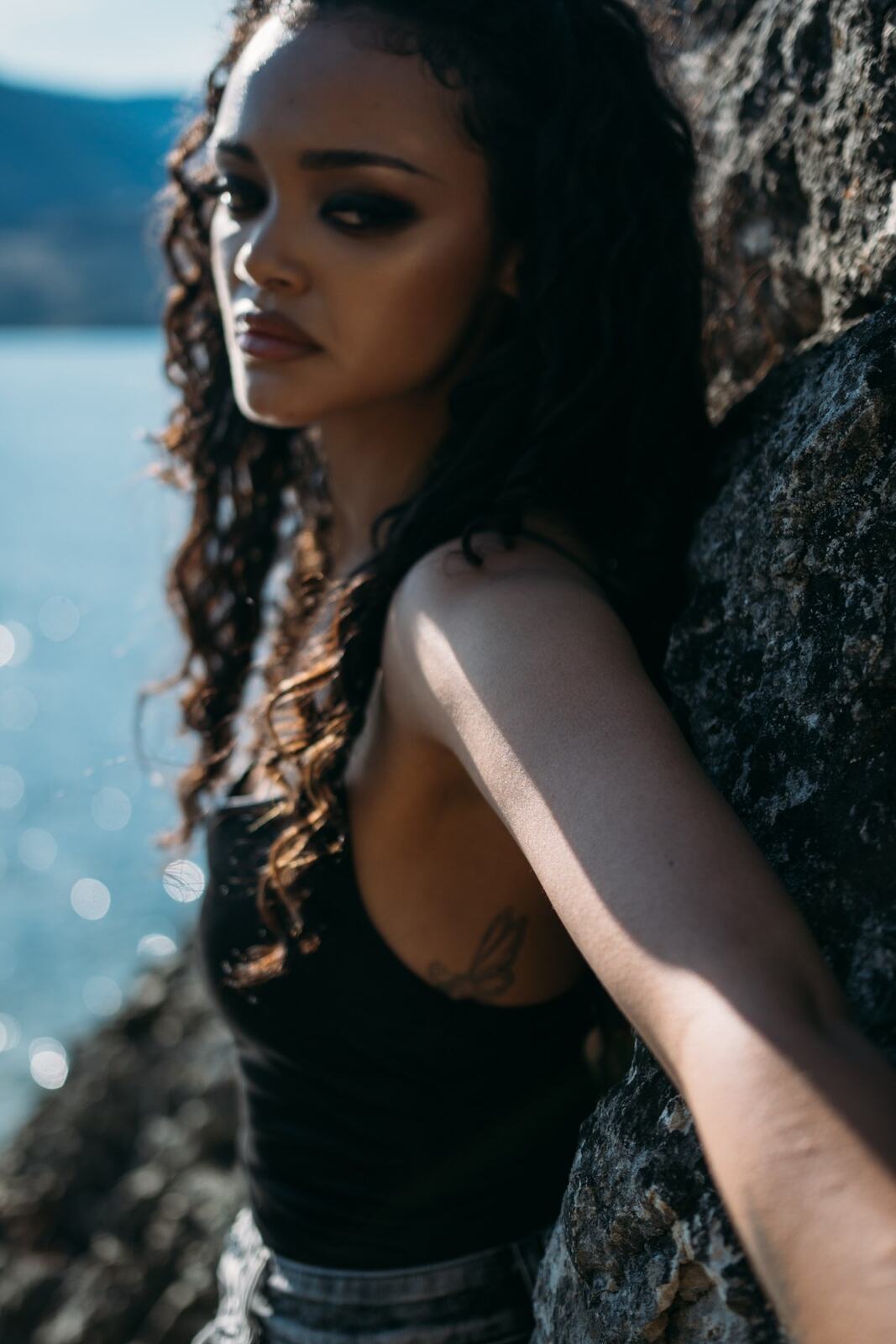 Alex by the Lake portraits story frame 2: Okanagan Lake portraits in Kelowna with black styling, rock, water, wind, direct expression and a cinematic beauty-portrait edge.