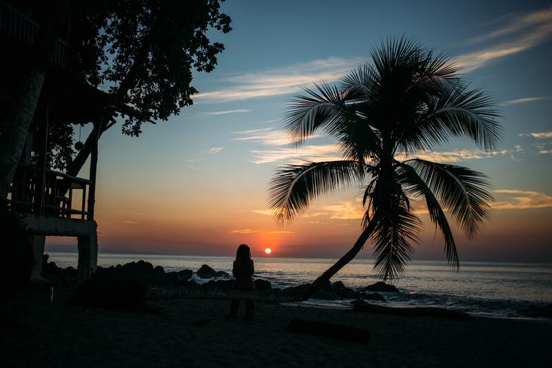 A palm, dark beach house and seated figure hold a deeply Costa Rican sunset over the Pacific. print option