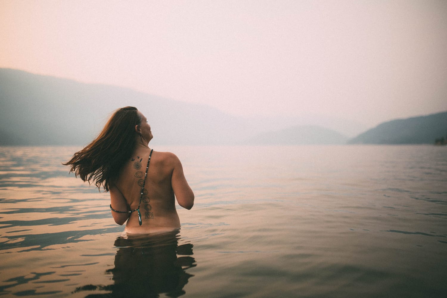 Erin at Mara Lake portraits story frame 8: Mara Lake portraits for Erin with driftwood, warm water, tattoo detail and soft evening light across the Shuswap shoreline.