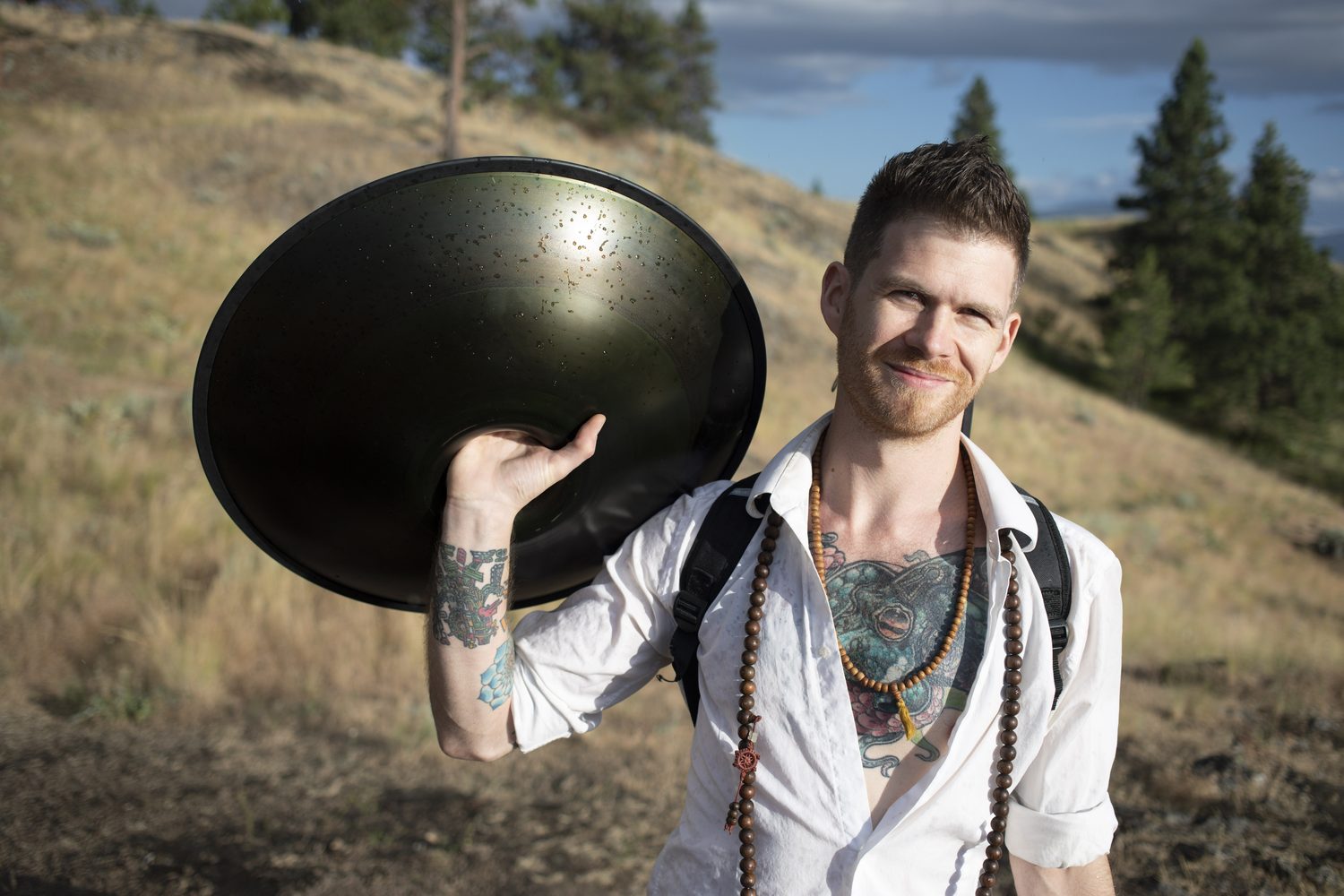 Jacob smiling beside a handpan with the lake and storm clouds behind him.