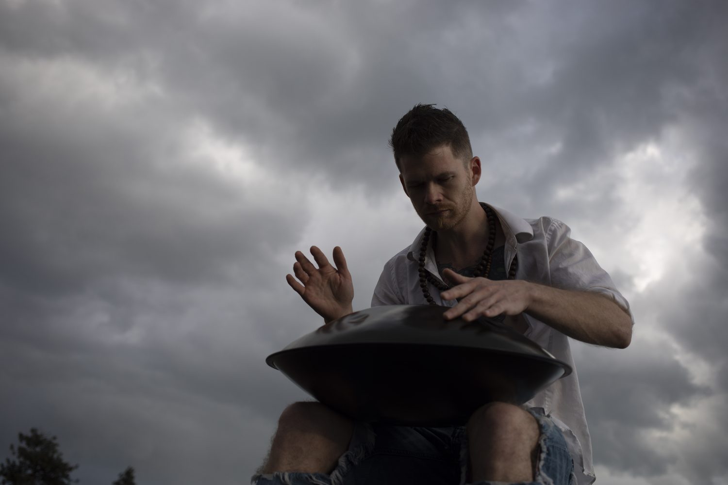 A low-angle portrait of Jacob playing handpan beneath dark clouds.