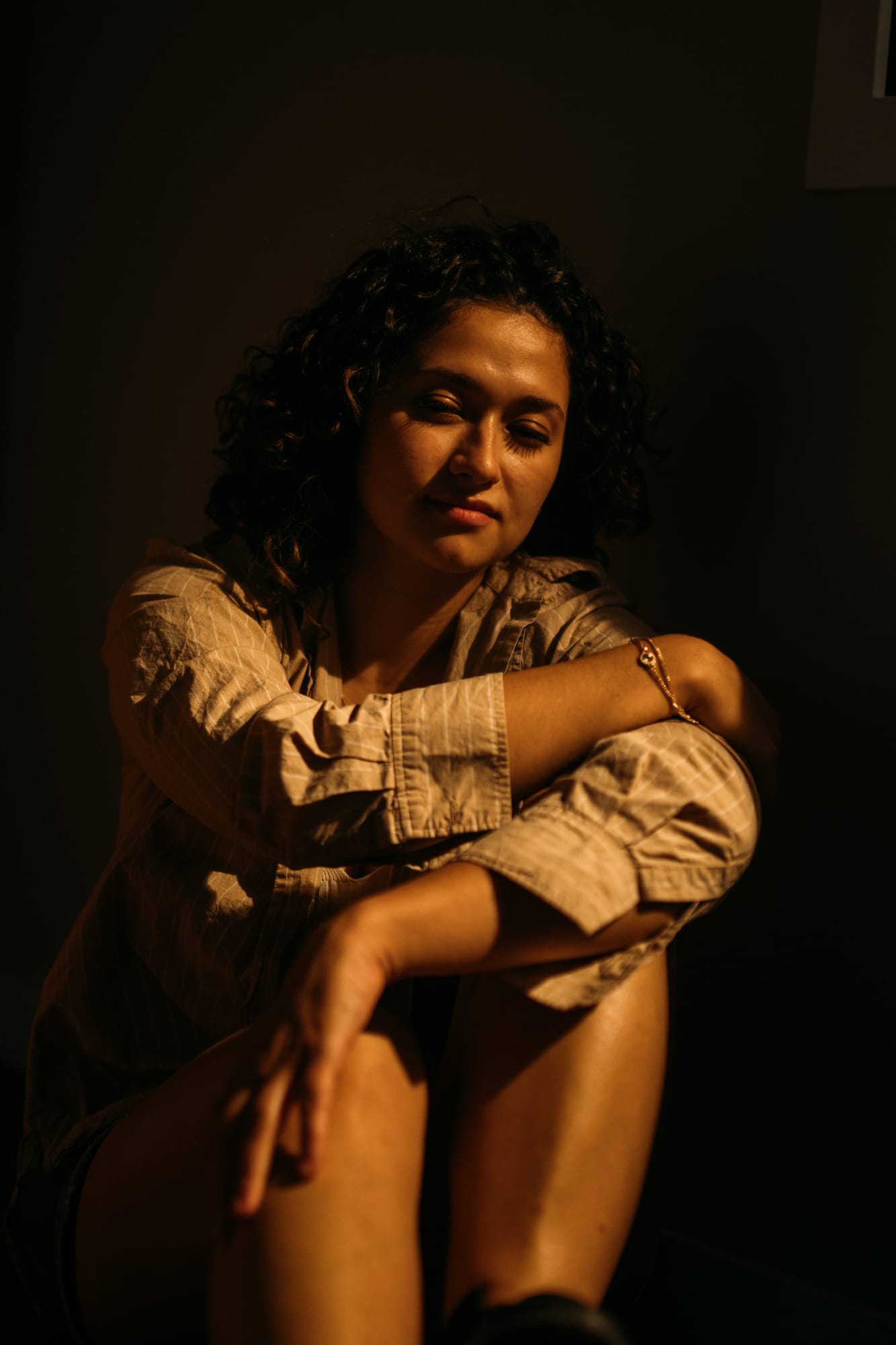 Marina in Warm Window Light portraits story frame 10: Marina in warm indoor light, with amber shadow, denim, black robe, green styling and an intimate editorial portrait mood.