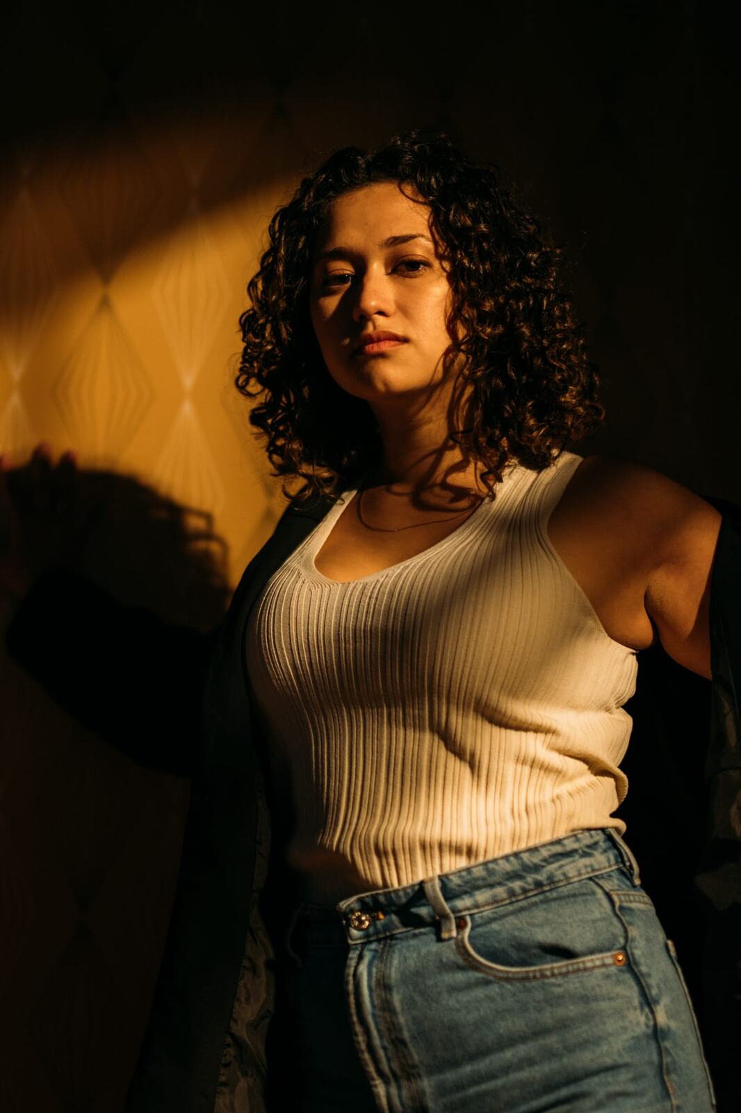 Marina in Warm Window Light portraits story frame 5: Marina in warm indoor light, with amber shadow, denim, black robe, green styling and an intimate editorial portrait mood.