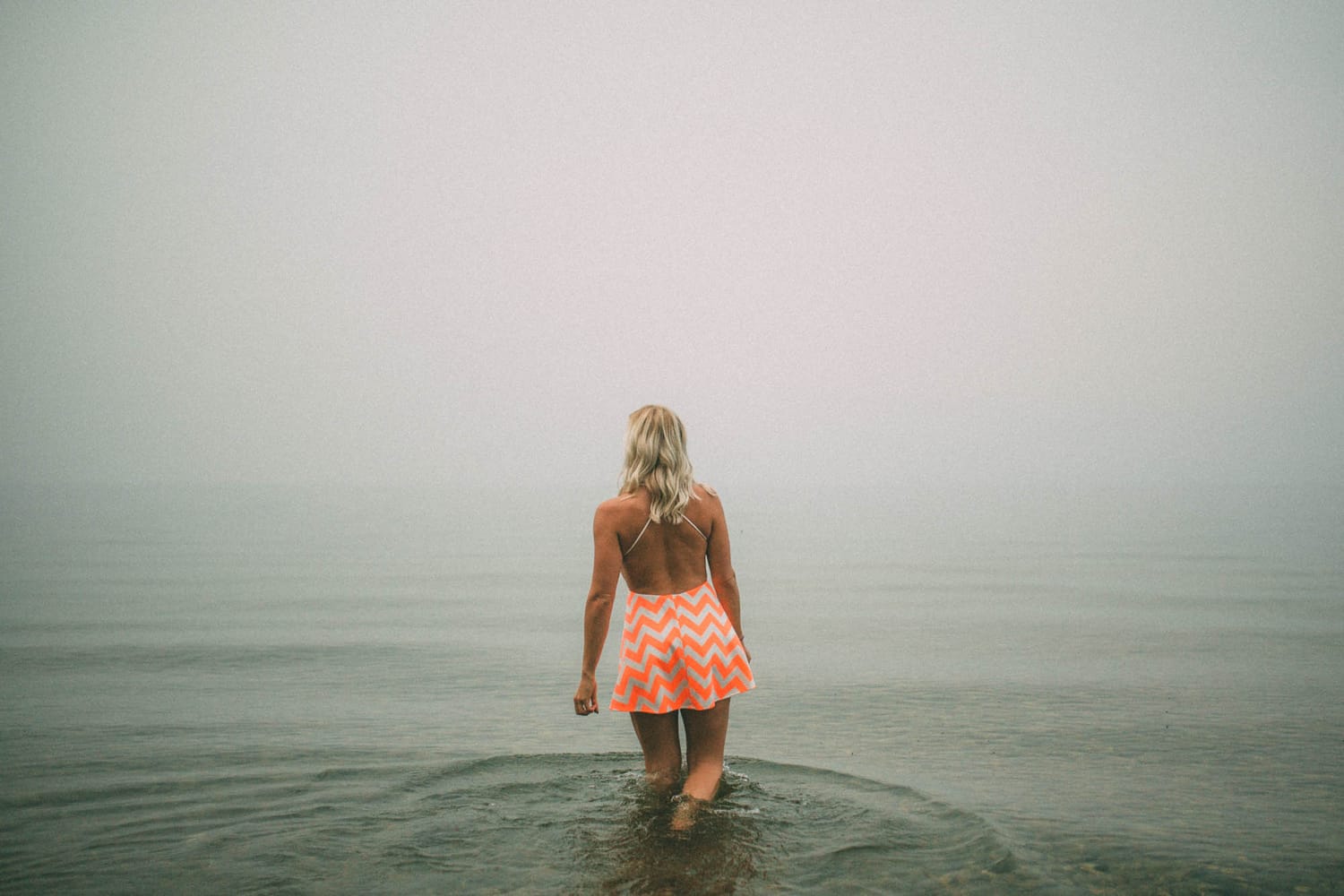 Megan Shallow portraits story frame 1: A fog-soft Kelowna lake portrait session for Megan Shallow, with water, orange dress movement, close body-language frames and quiet birds cutting through the mist.