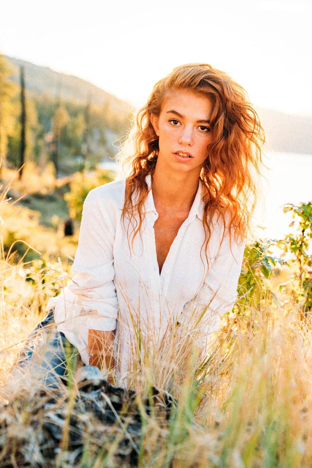 Mickie portraits story frame 6: A Kelowna portrait session for Mickie moving from golden grass and warm hillside light into lake-edge rocks, wind, white shirt styling and close editorial portraits.