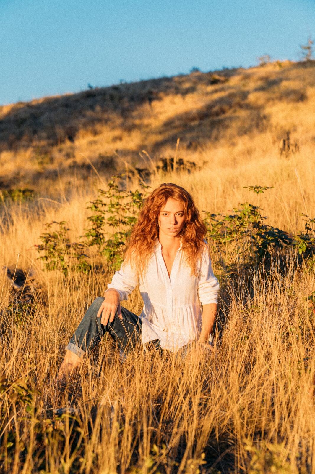 Mickie portraits story frame 7: A Kelowna portrait session for Mickie moving from golden grass and warm hillside light into lake-edge rocks, wind, white shirt styling and close editorial portraits.