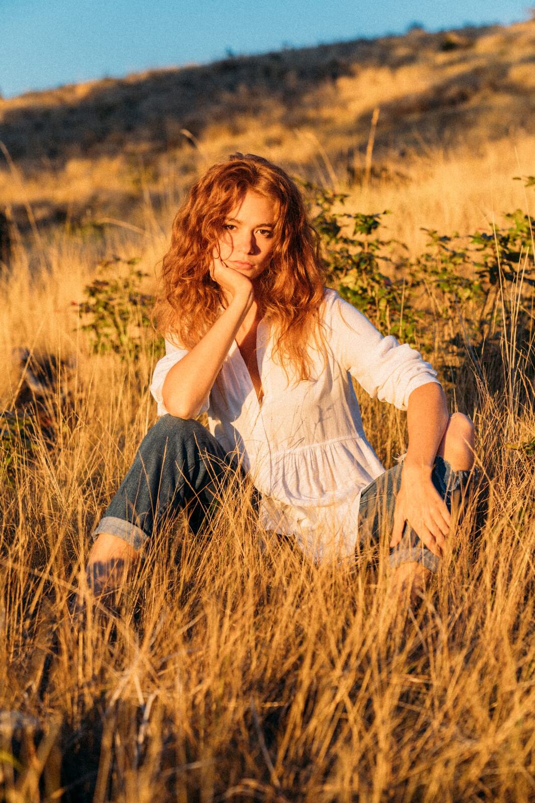 Mickie portraits story frame 8: A Kelowna portrait session for Mickie moving from golden grass and warm hillside light into lake-edge rocks, wind, white shirt styling and close editorial portraits.